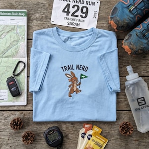 May include: A light blue t-shirt with the words "Trail Nerd" and a cartoon rabbit holding a flag. The image includes running shoes, a water bottle, a race bib, a map, car keys, a timer, and energy chews, all on a wooden surface.