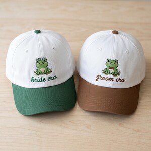 May include: Two baseball caps with white crowns and coloured brims. One has a green brim and the text "bride era" with a frog design. The other has a brown brim and the text "groom era" with a frog design. Both caps have a button on top.