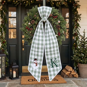 May include: A green and white gingham fabric ribbon hanging from a circular evergreen wreath. The ribbon features a white lace trim, a printed snowman on the left tail, and a large green letter K surrounded by foliage on the right tail.