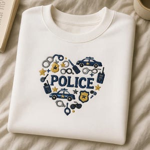May include: Cream-coloured sweatshirt featuring a heart-shaped embroidery of police symbols in navy and gold. The word "POLICE" is in the centre. Includes police cars, handcuffs, stars, and badges.