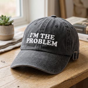 May include: A charcoal grey baseball cap with a distressed finish featuring the white embroidered text I'M THE PROBLEM on the front panel. The cap includes a curved brim, stitched eyelets, and a silver-toned metal adjustable buckle strap.