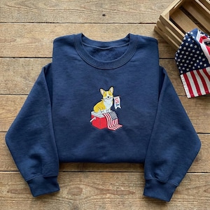 Pode incluir: Camisola azul marinho com um desenho bordado de um cão corgi sentado numa geleira vermelha, com uma bandeira americana e uma faixa com a inscrição "USA". A camisola está dobrada sobre uma superfície de madeira.