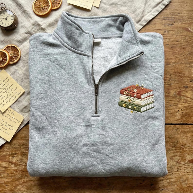 May include: A gray quarter-zip sweatshirt with a small embroidered design of stacked books with floral accents. The sweatshirt is folded neatly, with the zipper partially open. The background includes dried orange slices and vintage ephemera.