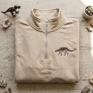 Pode incluir: Camisola bege com fecho de correr de um quarto de gola e um design bordado detalhado de um esqueleto de Anquilossauro. A camisola tem uma gola alta e um fecho de correr prateado. O gráfico do dinossauro é em tons de castanho e preto.