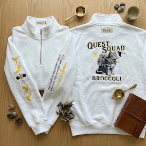 Könnte beinhalten: Weißes Sweatshirt mit Viertelreißverschluss, bestickt mit goldenen Drachen und Text auf dem Ärmel. Auf der Rückseite befindet sich eine Katze mit Rucksack und den Worten "QUEST SQUAD BROCCOLI". Ein Buch und eine Drachenfigur sind ebenfalls zu sehen.