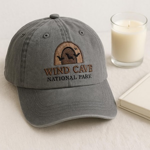 Peut inclure: Casquette de baseball grise avec le texte "Wind Cave National Park" brodé en marron. La casquette a une visière incurvée et un petit bouton rond sur le dessus. Une bougie dans un verre et un petit livre sont également dans l'image.