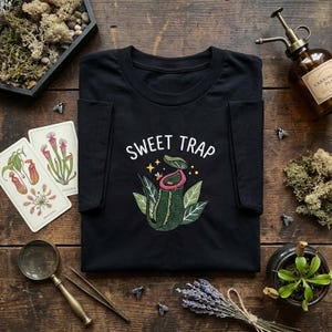 May include: A black short-sleeve t-shirt featuring an embroidered graphic of a green pitcher plant with a pink rim, surrounded by small yellow stars and a bee. The text SWEET TRAP is embroidered in white above the plant design on the chest.