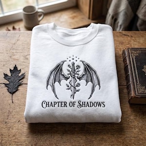 May include: A white crewneck sweatshirt featuring a black embroidered graphic of a sword entwined with roses and flanked by two bat-like wings. Above the sword are small stars, and below the design is the text "CHAPTER OF SHADOWS" in a serif font.