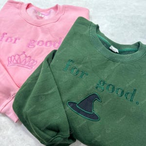 May include: Two sweatshirts, one pink and one green. The pink sweatshirt has the embroidered text "for good" and a crown. The green sweatshirt has the embroidered text "for good" and a witch's hat.