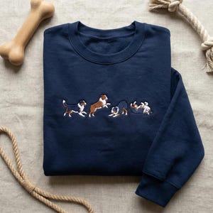 May include: Navy blue sweatshirt with embroidered dogs in various playful poses. The dogs are in shades of brown, black, and white. A wooden dog bone and rope toy are in the background.