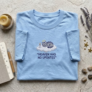 May include: A light blue t-shirt featuring an embroidered graphic of a grey tabby cat with a halo sleeping on a white cloud next to a lit candle. Below the graphic, the text reads "HEAVEN HAS NO UPDATES" in dark blue capital letters.
