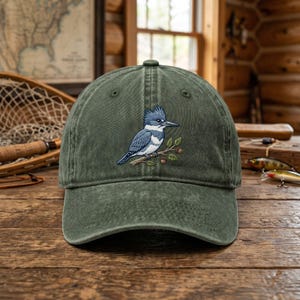 Könnte beinhalten: Grüne Baseballkappe mit gesticktem Bild eines blau-weißen Vogels auf einem Ast. Die Kappe besteht aus einem abgenutzten, strukturierten Stoff.