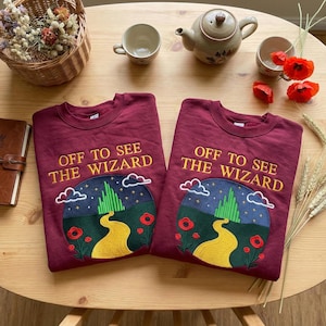 May include: Two maroon sweatshirts with the embroidered phrase "OFF TO SEE THE WIZARD". Each sweatshirt features a circular design with a yellow brick road, green Emerald City, and red poppies. The sweatshirts are on a wooden table.