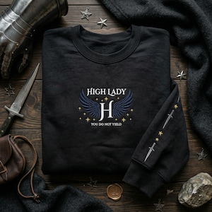 Peut inclure: Sweat-shirt noir avec l'inscription "High Lady" surmontant un "H" ailé et l'inscription "You Do Not Yield". La manche est ornée d'épées et d'étoiles. Le sweat-shirt est posé sur une surface en bois.