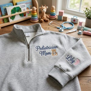 May include: A light grey quarter-zip sweatshirt. It features the embroidered text "Pediatrician Mom" and a teddy bear with a stethoscope. The sleeve has the name "Lily" and a pink footprint.