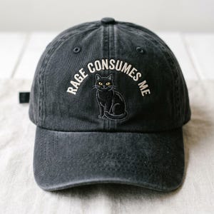 May include: A black baseball cap with the embroidered text "RAGE CONSUMES ME" above a black cat illustration with yellow eyes. The cap has a curved brim and a button on top.