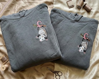 May include: Two gray hoodies with embroidered opossum designs. Each opossum is hanging from a branch with a pink tail and green leaves. The hoodies have drawstrings and are displayed on a beige surface.
