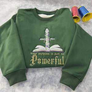 May include: A green sweatshirt with an embroidered design featuring a sword entwined with flowers atop an open book. The text "Your purpose is just as Powerful" is below the book. Two spools of thread are in the background.
