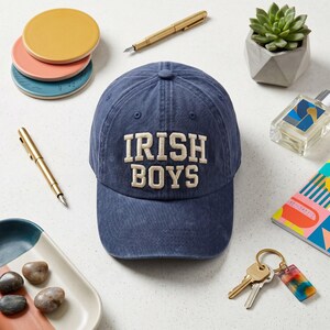 Pode incluir: Boné de beisebol azul marinho com o texto "IRISH BOYS" bordado em creme. A imagem também inclui uma suculenta em um vaso geométrico, canetas, porta-copos e um chaveiro.
