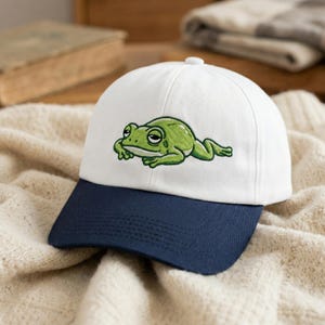 May include: A white baseball cap with a navy blue brim. The cap features an embroidered green frog design on the front. The frog has a relaxed expression. The cap is sitting on a cream-colored knitted blanket.