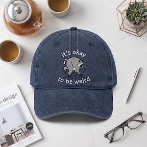 Könnte beinhalten: Eine marineblaue Baseballkappe mit verwaschener Textur. Auf der Vorderseite befindet sich eine gestickte Grafik eines grauen Waschbären, der sich bückt, sowie der weiß gestickte Text, it's okay to be weird.