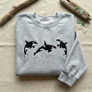 Könnte beinhalten: Ein hellgraues Sweatshirt mit Rundhalsausschnitt, bestickt mit drei schwarz-weißen Orca-Walen. Die Wale sind über die Vorderseite des Sweatshirts angeordnet. Das Kleidungsstück ist sauber auf einer neutralen Oberfläche gefaltet.
