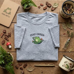 Peut inclure: Un t-shirt gris chiné avec l'inscription brodée "STAY GREEN" et une illustration de grenouille. L'image comprend également un carnet avec un dessin de grenouille et les mots "FROG NOTES". D'autres articles sont visibles, comme une tasse, un stylo et des plantes décoratives.