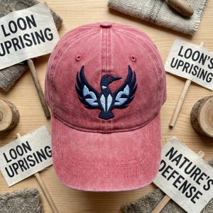 Könnte beinhalten: Verblasste rote Baseballkappe mit einem detaillierten, gestickten Eistaucher-Design in Marineblau und Hellblau. Auf kleinen Schildern steht der Text "LOON UPRISING". Ein modisches Accessoire für Outdoor-Aktivitäten.