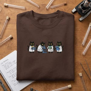 Peut inclure: Un sweat-shirt à col rond marron foncé orné d'un motif représentant quatre chats noirs vêtus de blouses de laboratoire blanches. Les chats effectuent des tâches scientifiques, utilisant un microscope, tenant des tubes à essai et un presse-papiers LAB RESULTS.