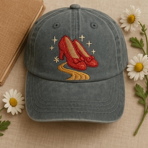 May include: A teal denim baseball cap with an embroidered design. The design features red high-heeled shoes, a yellow path, and gold stars. This whimsical design is centered on the front of the cap, offering a stylish accessory.