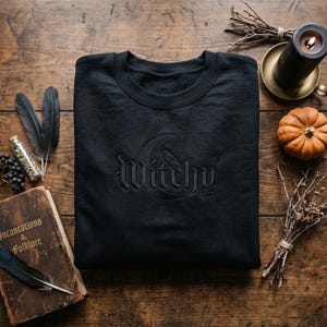 May include: A black crewneck sweatshirt featuring tonal black embroidery on the chest. The embroidery depicts a crescent moon and a small bird perched above the word Witchy, written in a gothic-style font. The garment has long sleeves.