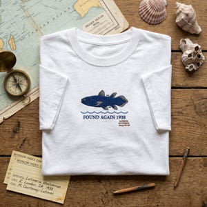 Pode incluir: Uma t-shirt branca de manga curta com um bordado de um celacanto azul-escuro sobre ondas azuis. O texto diz FOUND AGAIN 1938 e MUSEUM ACCESSION stamp #33.09. O design inclui pequenas etiquetas numéricas perto das barbatanas do peixe.