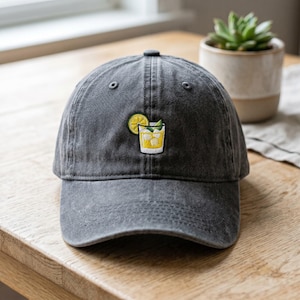 May include: A charcoal gray baseball cap with an embroidered cocktail design. The design features a yellow cocktail with a lemon slice and mint leaves. The cap is made of a soft, worn fabric.