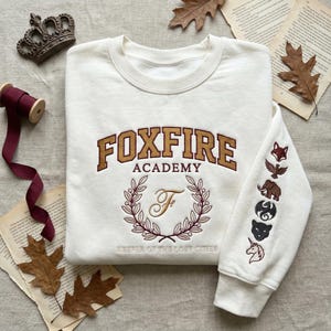 May include: Cream-colored sweatshirt with the text "FOXFIRE ACADEMY" in brown and gold lettering. The sleeve features embroidered animal and symbol designs. The sweatshirt also has a wreath design with the letter "F" and the text "KEEPER OF THE LOST CITIES".