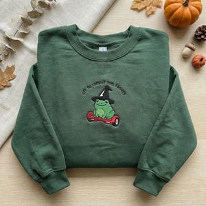 May include: Dark green sweatshirt featuring an embroidered design of a green frog in a witch hat on a red hoverboard. The text "off to commit tom foolery" is arched above the frog. The sweatshirt is displayed on a neutral surface with autumn decorations.