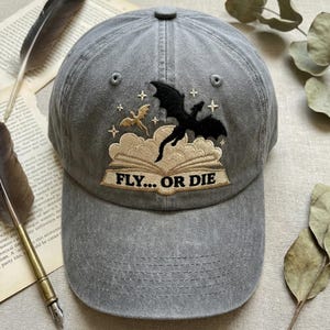 May include: A grey baseball cap featuring an embroidered design of an open book with clouds rising from the pages. A large black dragon and a smaller gold dragon are depicted above the book, accompanied by small star icons and the text FLY... OR DIE.