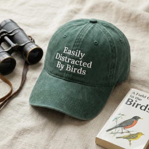 Peut inclure: Casquette de baseball verte avec le texte "Easily Distracted By Birds" en blanc. La casquette est à côté de jumelles et d'un livre intitulé "A Field Guide to the Birds". Les articles sont sur une surface de couleur neutre.
