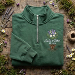 May include: A forest green quarter-zip sweatshirt with a floral embroidery design. The embroidery includes lavender, daisies, and greenery, with the names "Sophia & Ethan" and the date "12.2026" below. The sweatshirt is folded on a wooden surface.