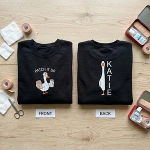 May include: Black sweatshirt with a cartoon goose wearing a nurse's hat, holding bandages and a cup, with the text "PATCH IT UP". The back of the sweatshirt has a goose design and the name "KATIE".