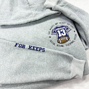 May include: A light gray hoodie with a hood and long sleeves. The left sleeve has the embroidered text "FOR KEEPS" in navy blue. The chest features a navy blue and white football jersey design with the name "KAVANAGH" and the number "13".