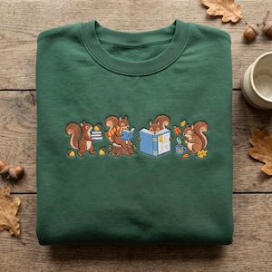 May include: A dark green crewneck sweatshirt featuring an embroidered graphic of four brown squirrels. One squirrel carries books, another sits on a stump reading, a third holds an open book, and the fourth sits beside a steaming blue mug.
