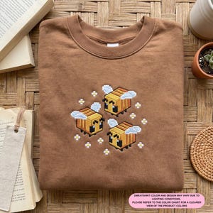 May include: A brown sweatshirt featuring an embroidered design of three pixelated bees with white cloud accents and small white flowers. The sweatshirt has a crew neck and a tag on the collar.