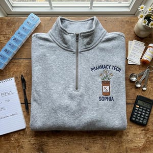 May include: A heather gray quarter-zip sweatshirt featuring an embroidered design on the left chest. The embroidery depicts a brown prescription bottle with white daisies and the text PHARMACY TECH above and the name SOPHIA below in navy blue.