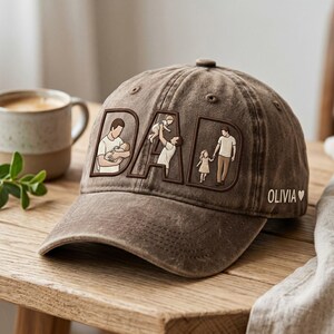 May include: A brown baseball cap with the word "DAD" embroidered on the front. The letters feature illustrations of a father with his children. The name "OLIVIA" is embroidered on the side, followed by a heart symbol. The cap is sitting on a wooden surface.