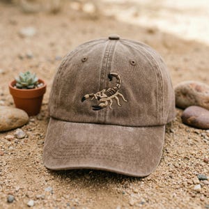 May include: A brown baseball cap with a distressed, washed-out finish. The front panel features a detailed, centred embroidery of a scorpion in shades of tan, brown, and black, showcasing its segmented body, pincers, and curved tail.
