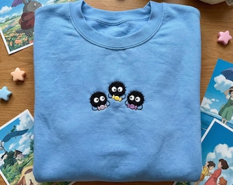 May include: A light blue sweatshirt featuring an embroidered design of three black soot sprites with colorful accents. The sweatshirt is surrounded by postcards and plush toys, creating a whimsical aesthetic. The design is centered on the chest.