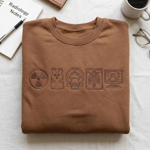 May include: A brown sweatshirt with a crew neck and medical imaging symbols. The symbols include a radiation symbol, a vest, an MRI machine, a rib cage, and a computer screen. The words "Radiology Notes" are visible on a notepad.