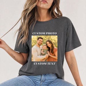 Op de afbeelding: Donkergrijs t-shirt met een familiefoto en de tekst "CUSTOM PHOTO" boven de afbeelding en "CUSTOM TEXT" eronder. De foto toont een lachend gezin van drie personen. Het shirt heeft een casual stijl.