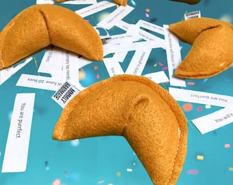 May include: A close-up of a fortune cookie with a light brown color. The cookie is made of felt and has a white seam around the edges. The cookie is surrounded by confetti and fortune cookie slips with various messages. The slips have the text "You are purrfect", "You will have 10 lives", "empty lap needs", "outerbox will always be", "climb to greatness", and "You are".