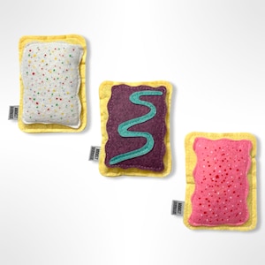 Puede incluir: Tres versiones de juguete de fieltro de un popular desayuno. La primera es una pastelería blanca con chispas de colores. La segunda es una pastelería morada con un remolino verde. La tercera es una pastelería rosa con chispas blancas. Cada pastelería tiene una corteza amarilla y el texto "MAKEY MAKEY" en la parte inferior.